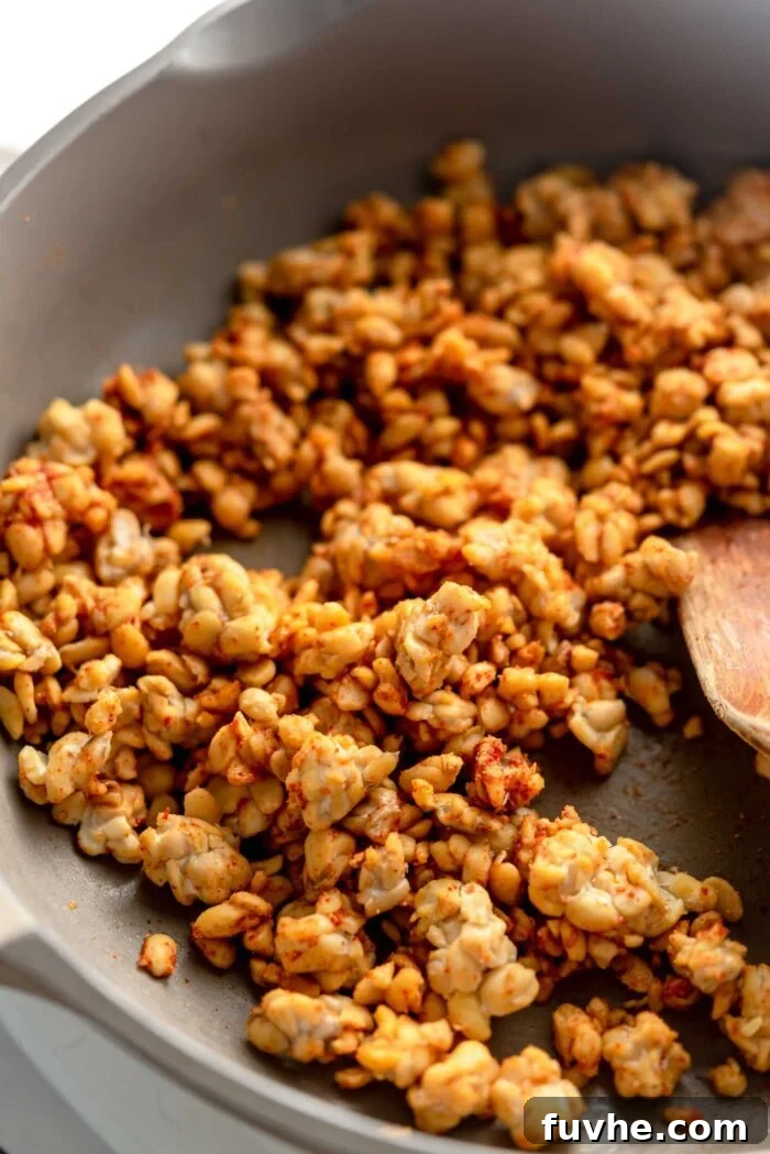 Crumbled crispy tempeh bits with paprika and garlic powder cooking in a skillet with a wooden spoon.