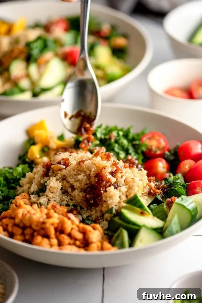 Drizzling maple chili dressing from a spoon over a grain bowl with tempeh, tomato, avocado and cucumber.