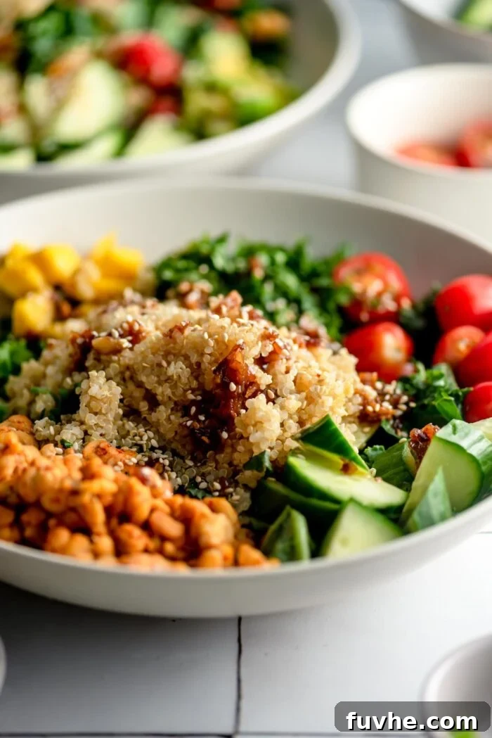 A healthy vegetarian quinoa grain bowl with avocado, mango, tempeh, cucumber, cilantro, tomato, green onion and shallot garlic dressing. A second grain bowl is in the background.