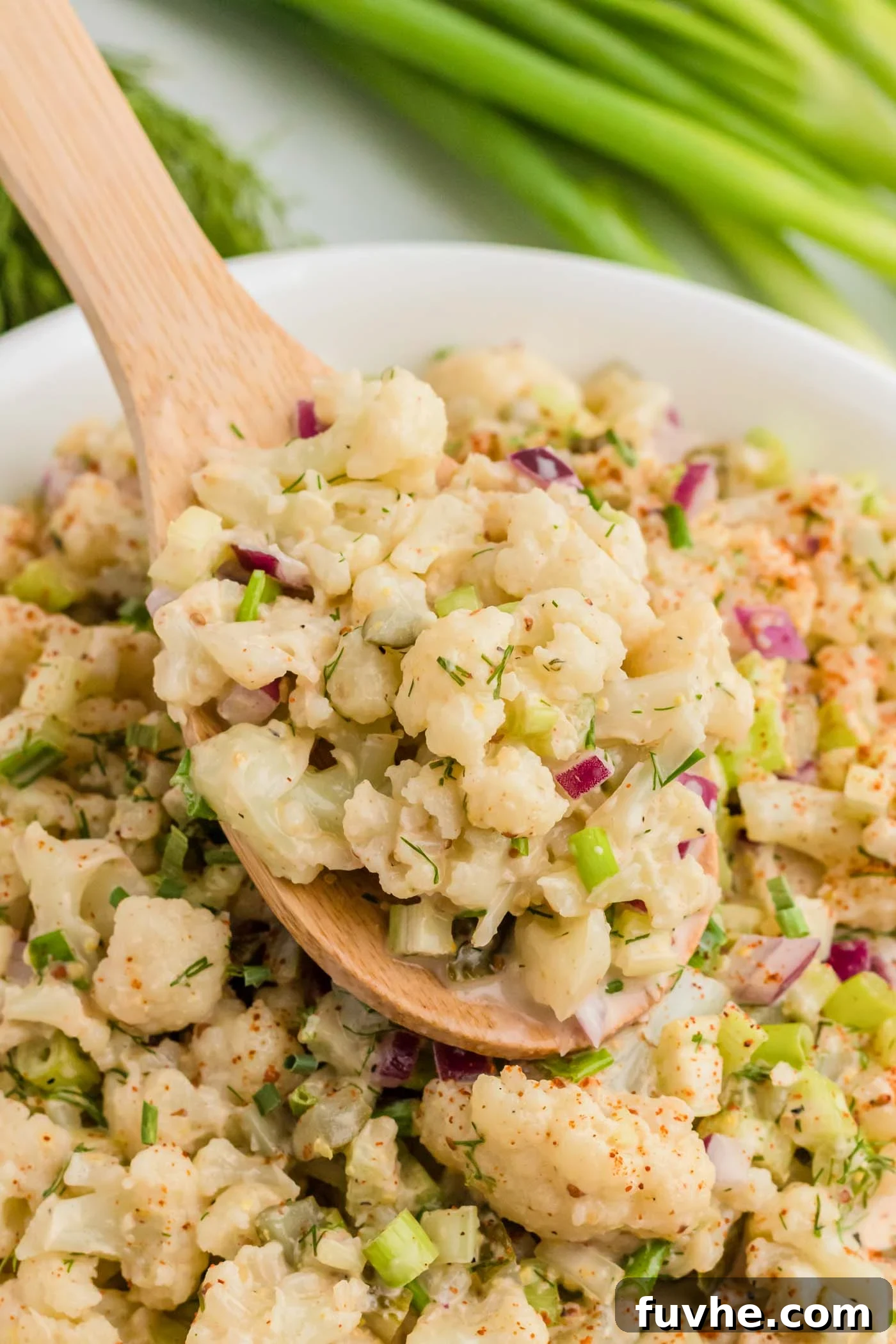 A wooden spoon scoops a generous portion of vegan cauliflower mock potato salad from a large serving bowl, highlighting the creamy texture and vibrant green specks of chives. In the background, fresh green onions hint at the salad's refreshing ingredients.