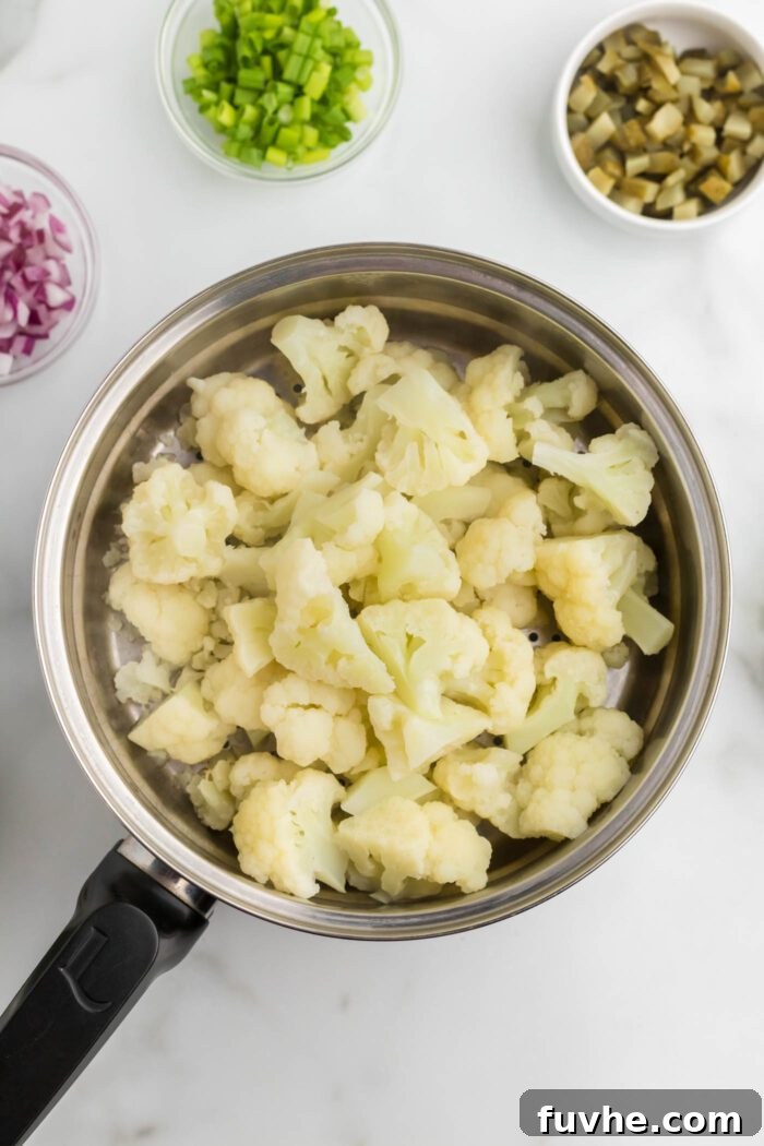 Perfectly steamed cauliflower florets rest in a steamer pot, ready for the next step. Beside the pot, three small bowls hold neatly diced dill pickles, vibrant red onion, and fresh chopped green onion, illustrating the prepared ingredients.