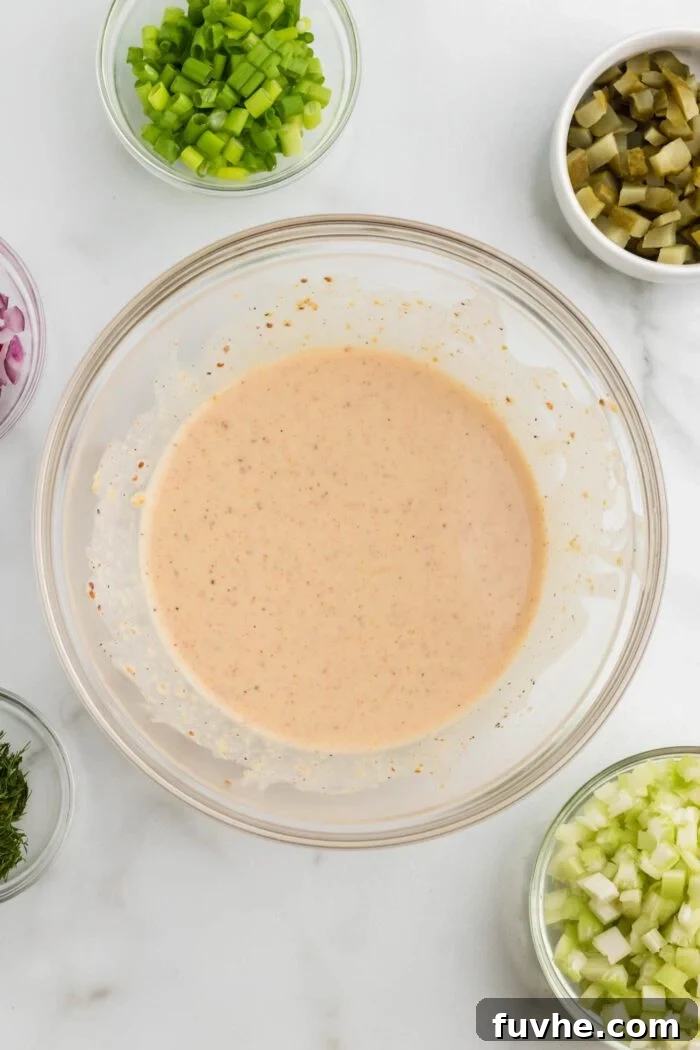 A smooth and creamy vegan potato salad dressing is mixed in a clear glass bowl with a whisk, surrounded by small bowls containing chopped celery, dill pickle, and scallions, all prepared and ready to be incorporated into the salad.