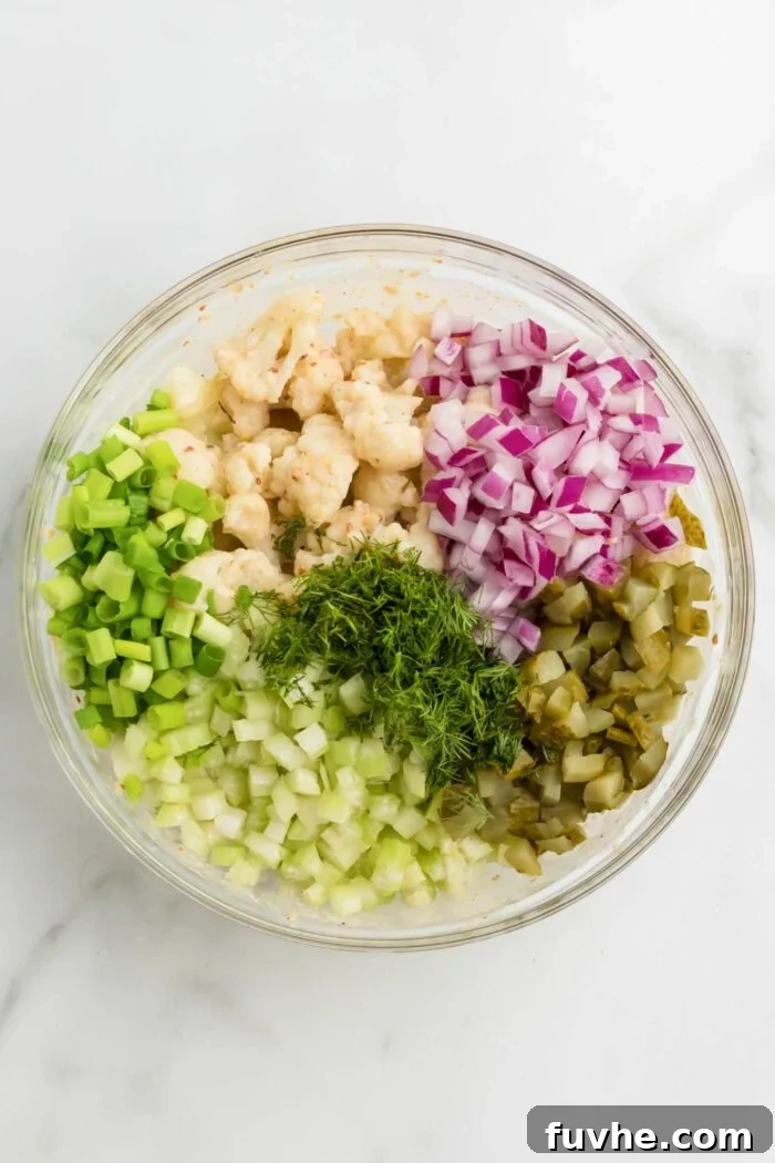Steamed cauliflower florets, vibrant red onion, crisp celery, tangy dill pickle, and fresh dill and scallions are harmoniously combined in a large glass mixing bowl on a sleek marble countertop, waiting to be tossed with the creamy dressing.