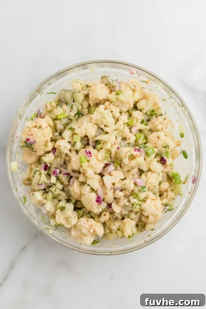 A mouth-watering image of a no-potato "potato" salad, expertly crafted with cauliflower, mixed together in a clear glass mixing bowl.