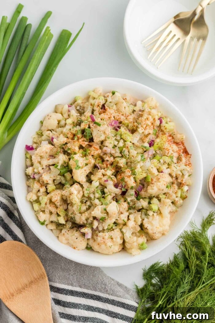 A stunning close-up of mock potato salad, made with tender cauliflower florets, crunchy dill pickle, and crisp celery, beautifully presented in a bowl and topped with fresh chives and a dusting of smoked paprika. Fresh green onions and dill sprigs artfully placed beside the bowl complete the inviting scene.