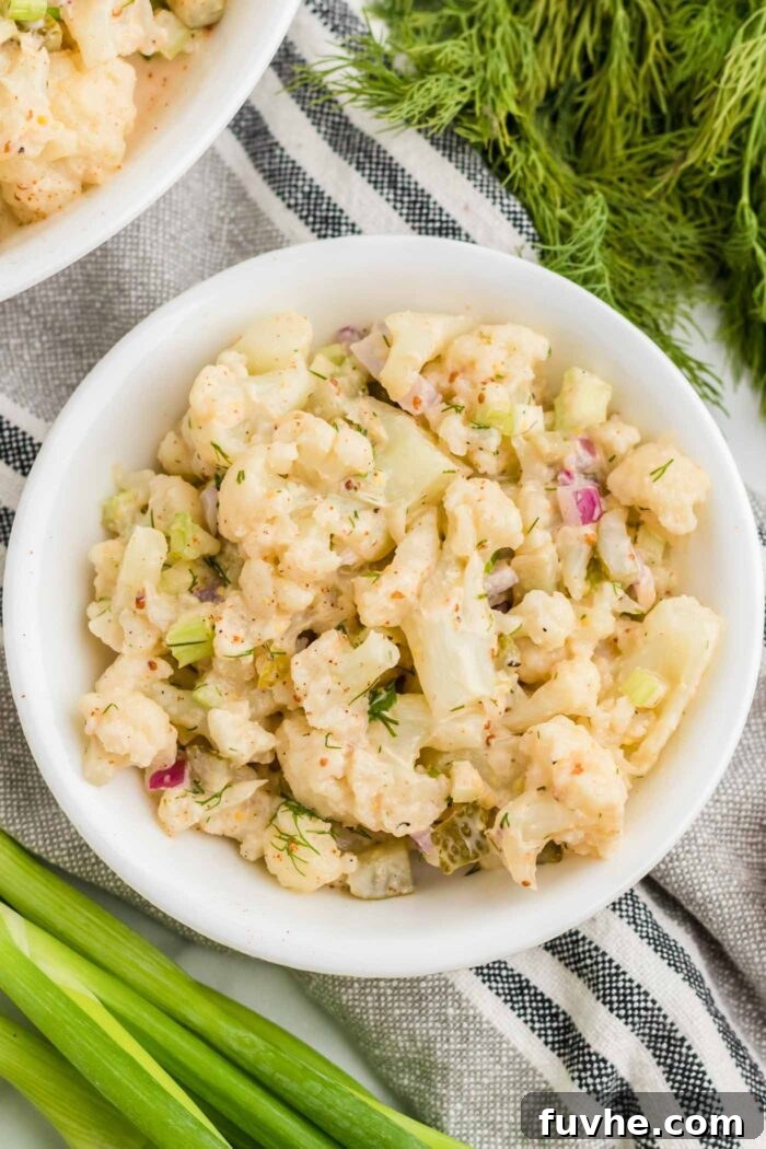 An inviting overhead view captures a generous bowl of vegan cauliflower potato salad, showcasing a delightful mix of tender cauliflower florets, crisp dill pickle bits, crunchy celery, and vibrant red onion. Sprinkled with a touch of smoked paprika and garnished with fresh dill and scallions, this dish is a feast for the eyes and the palate.