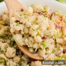 Wooden spoon scooping cauliflower mock potato salad from a large serving bowl of salad. There are some green onions in the background.