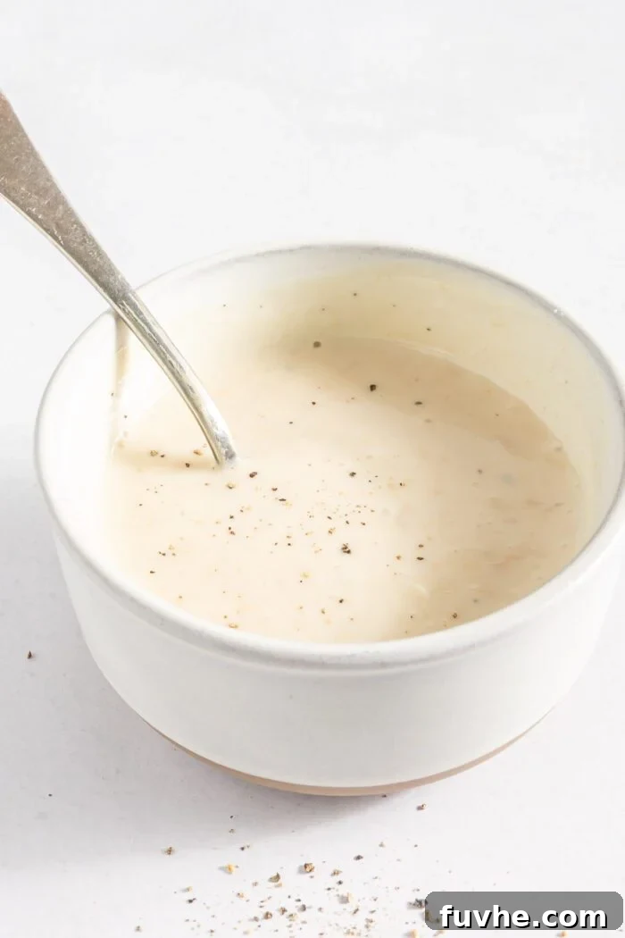 A small bowl filled with the creamy, mayo-based dressing for the broccoli salad, with a spoon resting in it, showcasing its smooth texture.