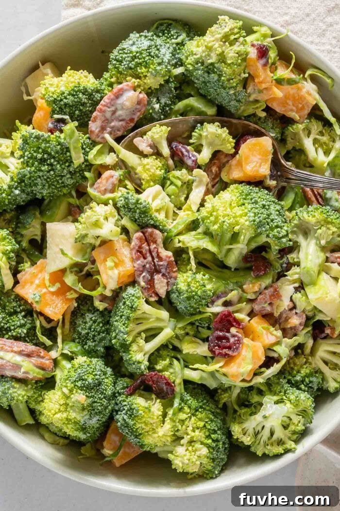 An overhead close-up view of a perfectly chilled bowl of broccoli apple salad, generously studded with pecans, cranberries, tender butternut squash, and shaved Brussels sprouts. A spoon waits patiently in the bowl.
