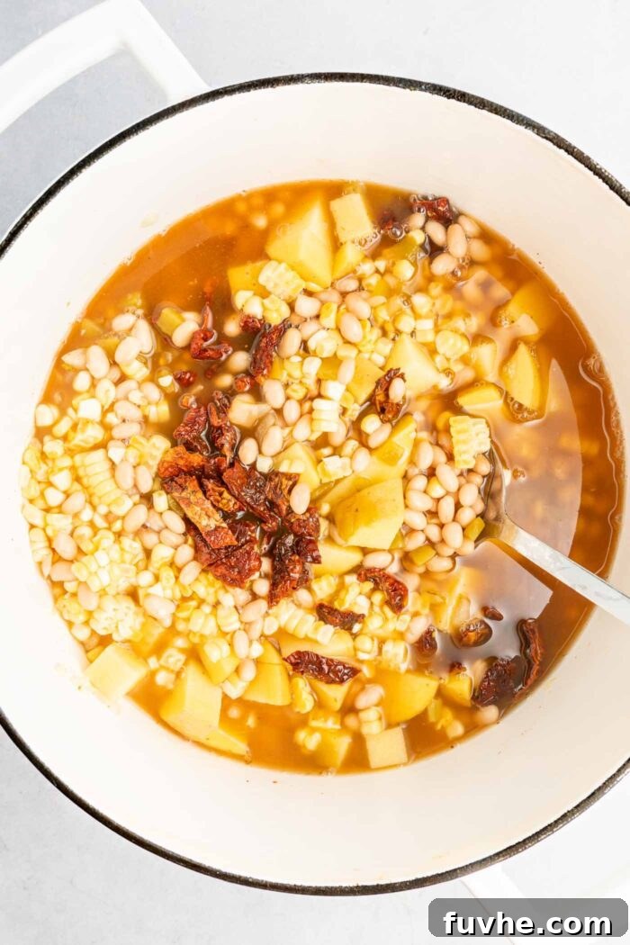 Sunny Vegan Corn Chowder 5 A large soup pot filled with a vibrant mixture of fresh corn kernels, chopped sun-dried tomatoes, diced potatoes, white beans, and vegetable broth, simmering gently on the stovetop.