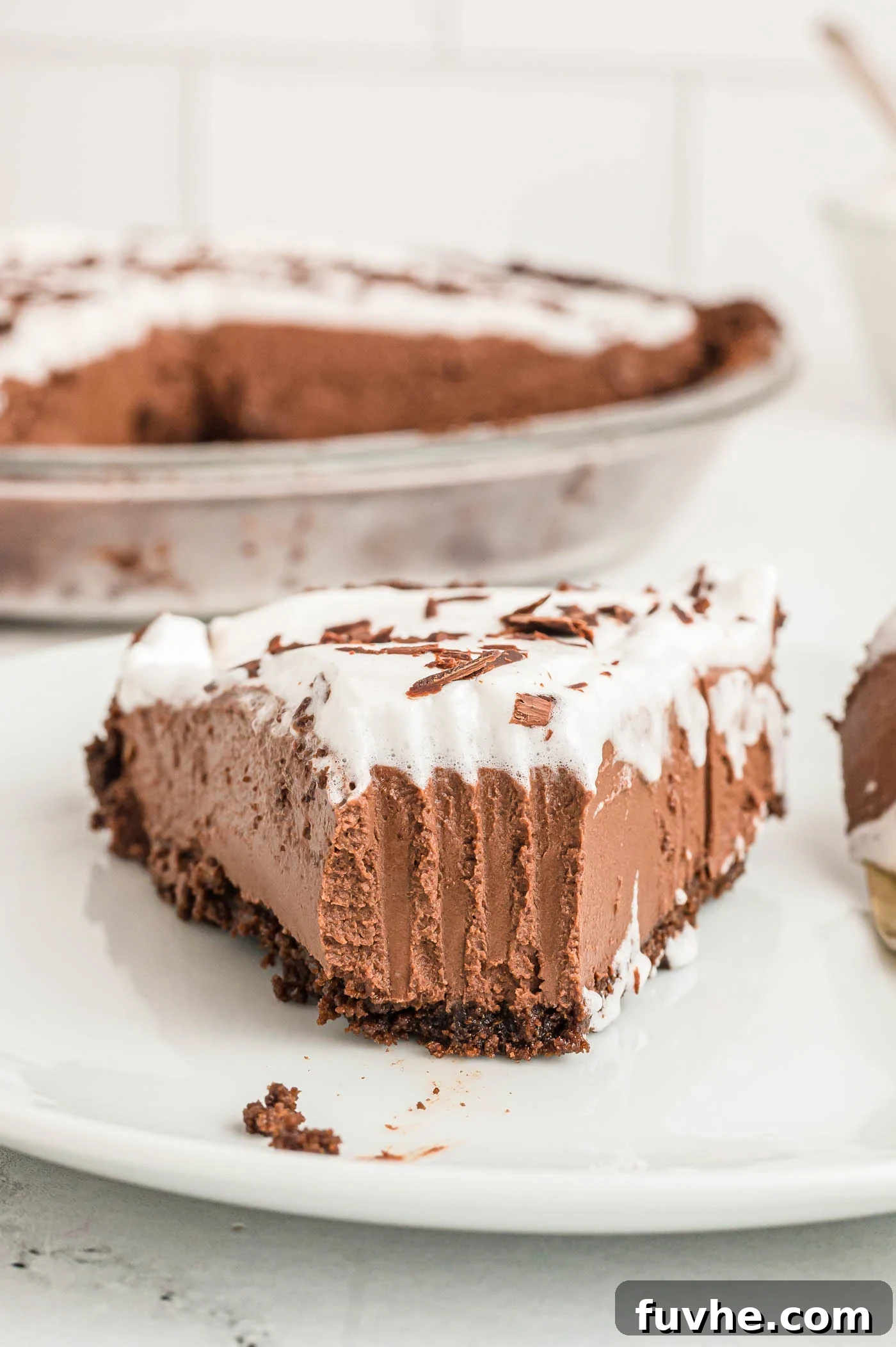 A perfectly sliced no-bake vegan chocolate pie with a rich chocolate crust, topped generously with fluffy whipped cream and delicate chocolate shavings. A bite has been taken, revealing the incredibly smooth, mousse-like interior of the pie. A fork rests beside the slice.
