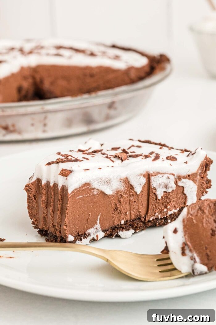 A beautifully presented slice of no-bake vegan chocolate pie, complete with chocolate crust, fluffy whipped cream, and chocolate shavings, resting on a small plate. A fork with a bite of pie is positioned on the plate, emphasizing the deliciousness.