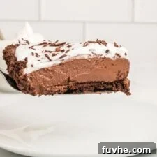A slice of vegan chocolate pie with a chocolate crust topped with whipped cream on a spatula held over a plate against a white subway tile background.