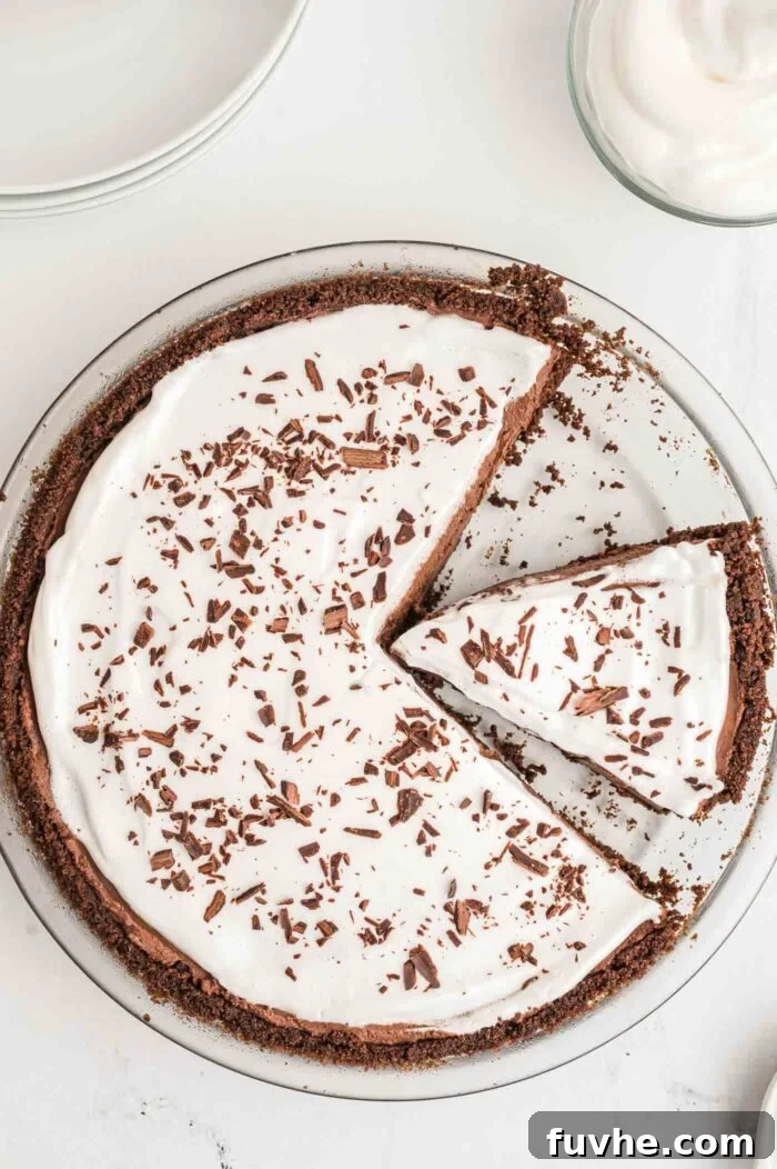 An overhead shot of a finished no-bake vegan chocolate pie, beautifully topped with generous swirls of whipped cream and delicate chocolate shavings. One slice has been removed, highlighting the inviting texture, and another slice is ready to be served.