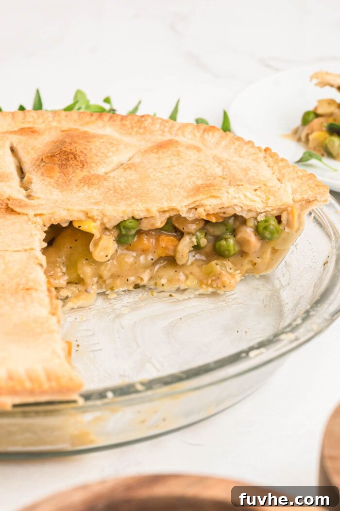 An inviting angled view into a glass pie dish showcasing a freshly baked vegan chickpea pot pie, from which a slice has been removed. The vibrant filling, rich with peas, carrots, corn, chickpeas, celery, and potato, is perfectly visible and steaming invitingly.