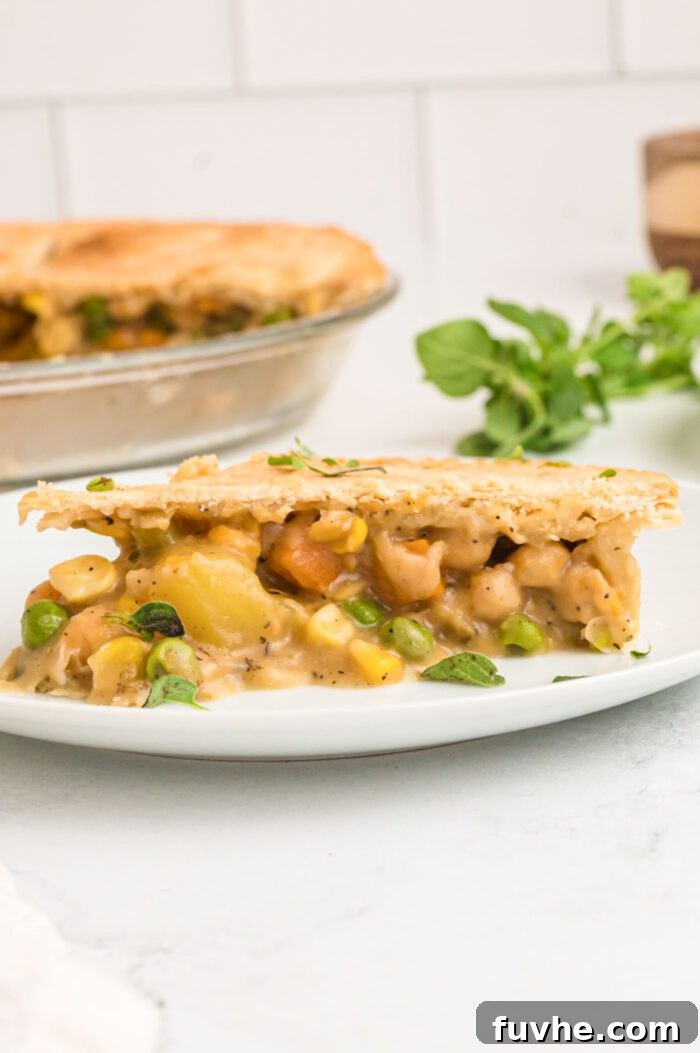 A delectable slice of vegan chickpea pot pie presented on a white plate, highlighting the rich and chunky filling of potatoes, carrots, peas, corn, and chickpeas. In the soft-focus background, the full golden-crusted pot pie rests in its glass dish, ready for more servings.
