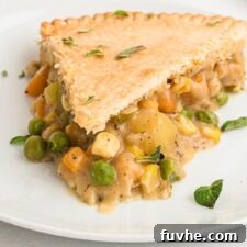 A slice of veggie pot pie with a potato, carrot, peas, corn and chickpea filling on a plate. The whole pie can be seen in the background.