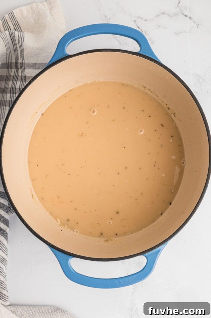 A smooth, thick, and creamy mixture of flour, butter, vegetable stock, and almond milk simmering in a large soup pot with handles, forming the luscious sauce for the pot pie.
