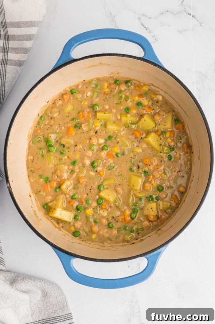 A thick and vibrant pot pie filling simmering in a large soup pot, featuring an array of colorful vegetables including diced potatoes, chickpeas, peas, corn, and carrots, all coated in a rich, creamy sauce.