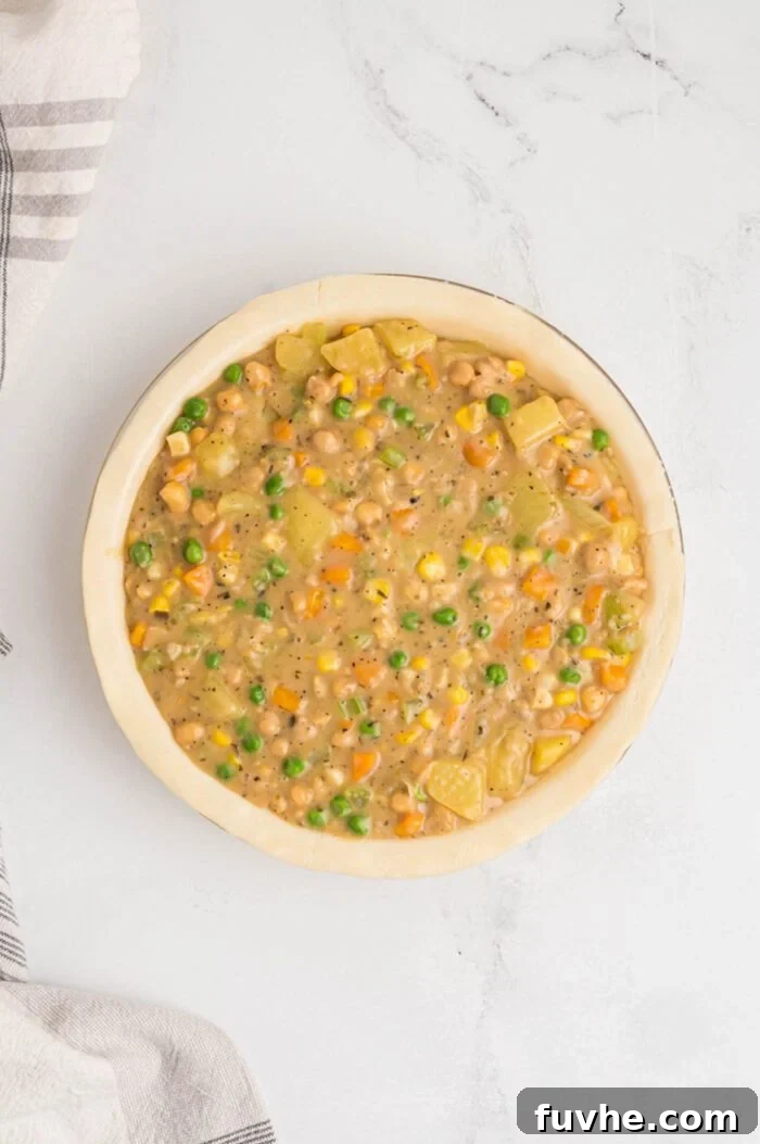 An unbaked pot pie filling, consisting of potatoes, chickpeas, carrots, corn, peas, and celery, generously spooned into a pie dish, ready for the top crust.