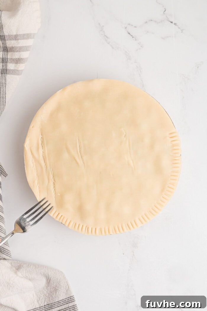 A close-up of a fork pressing along the edge of a pot pie to crimp and seal the top and bottom pie crusts together, creating a decorative and secure border.