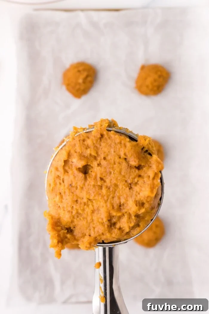 Pumpkin cookie dough in a cookie scoop held over a baking tray of raw cookies.