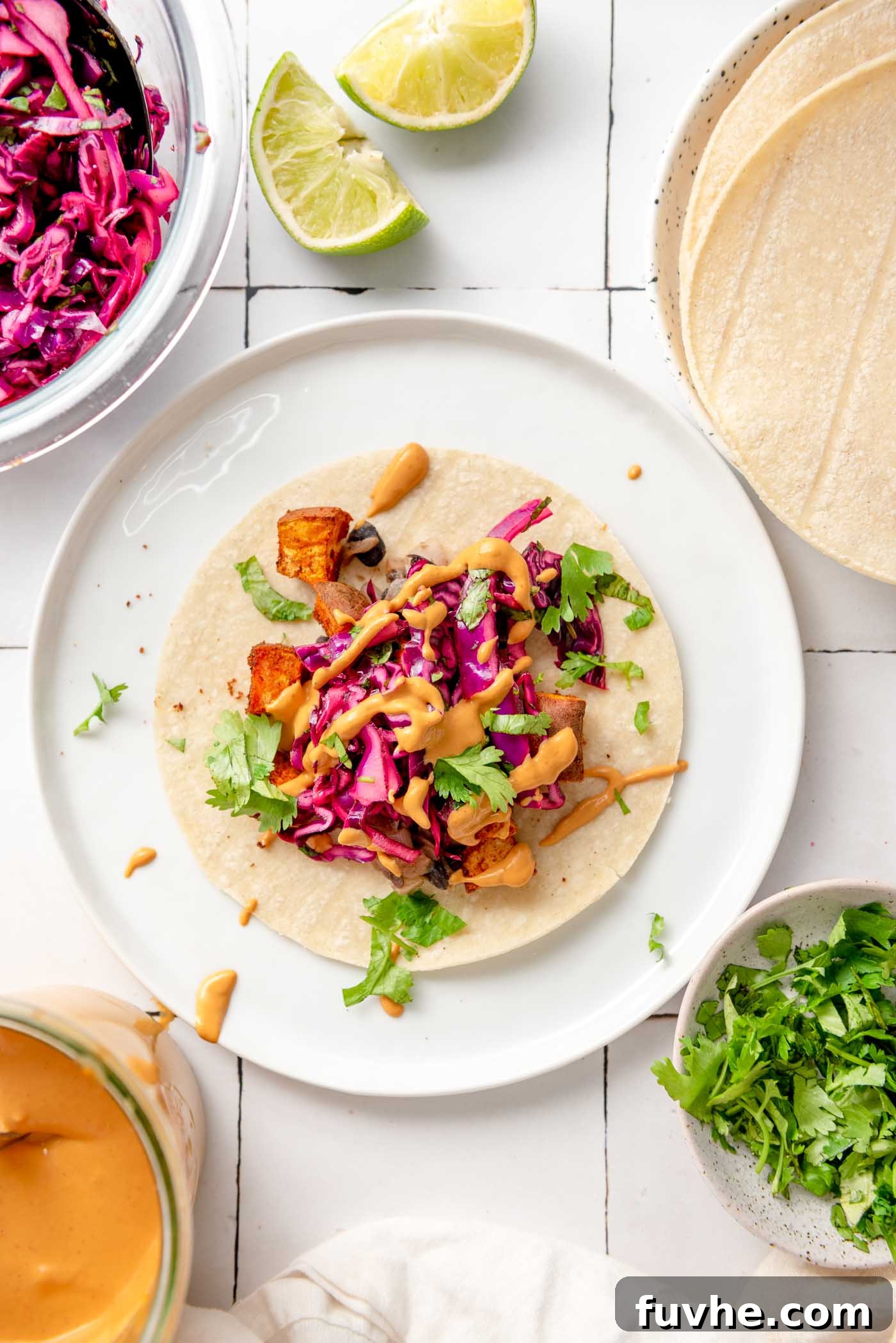 Spiced Sweet Potato and Black Bean Tacos 2 A vibrant sweet potato and black bean taco, generously topped with crisp red cabbage slaw and a swirl of creamy chipotle sauce. The plate also features a stack of warm tortillas, alongside small bowls of fresh cilantro, extra slaw, and the irresistible crema, all set for a delightful meal.