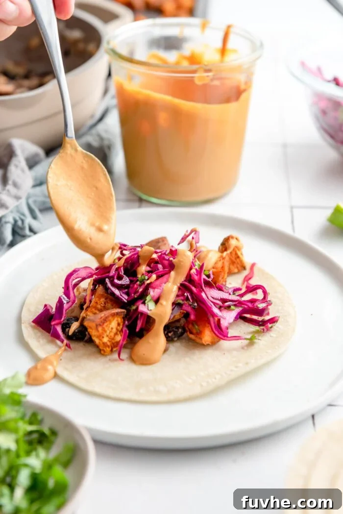 Spiced Sweet Potato and Black Bean Tacos 11 A spoon elegantly drizzles rich, creamy queso sauce over a perfectly assembled sweet potato and black bean taco on a plate, adding the finishing touch.