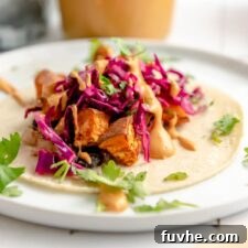 A vibrant and appetizing sweet potato and black bean taco, elegantly topped with crisp red cabbage slaw, fresh cilantro, and a swirl of creamy chipotle sauce. The background features a jar of the delicious queso, with a spoon inside, ready for more.