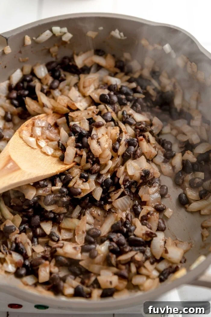 Spiced Sweet Potato and Black Bean Tacos 9 Hearty black beans and sautéed onions, infused with aromatic spices, cooking gently in a skillet, emitting a savory steam. A wooden spoon rests within the mixture, ready for stirring.