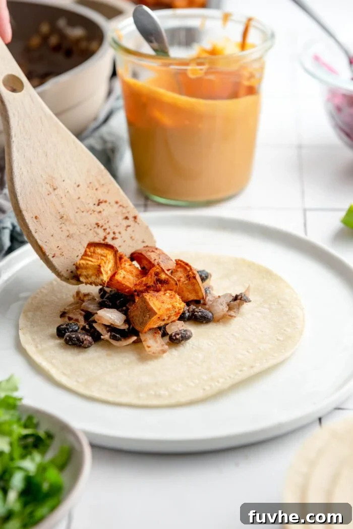 Spiced Sweet Potato and Black Bean Tacos 10 A warm corn tortilla on a plate, layered with a flavorful onion and black bean mixture. A wooden spoon carefully adds spiced sweet potato cubes, while a glass jar of creamy queso sauce awaits in the background.