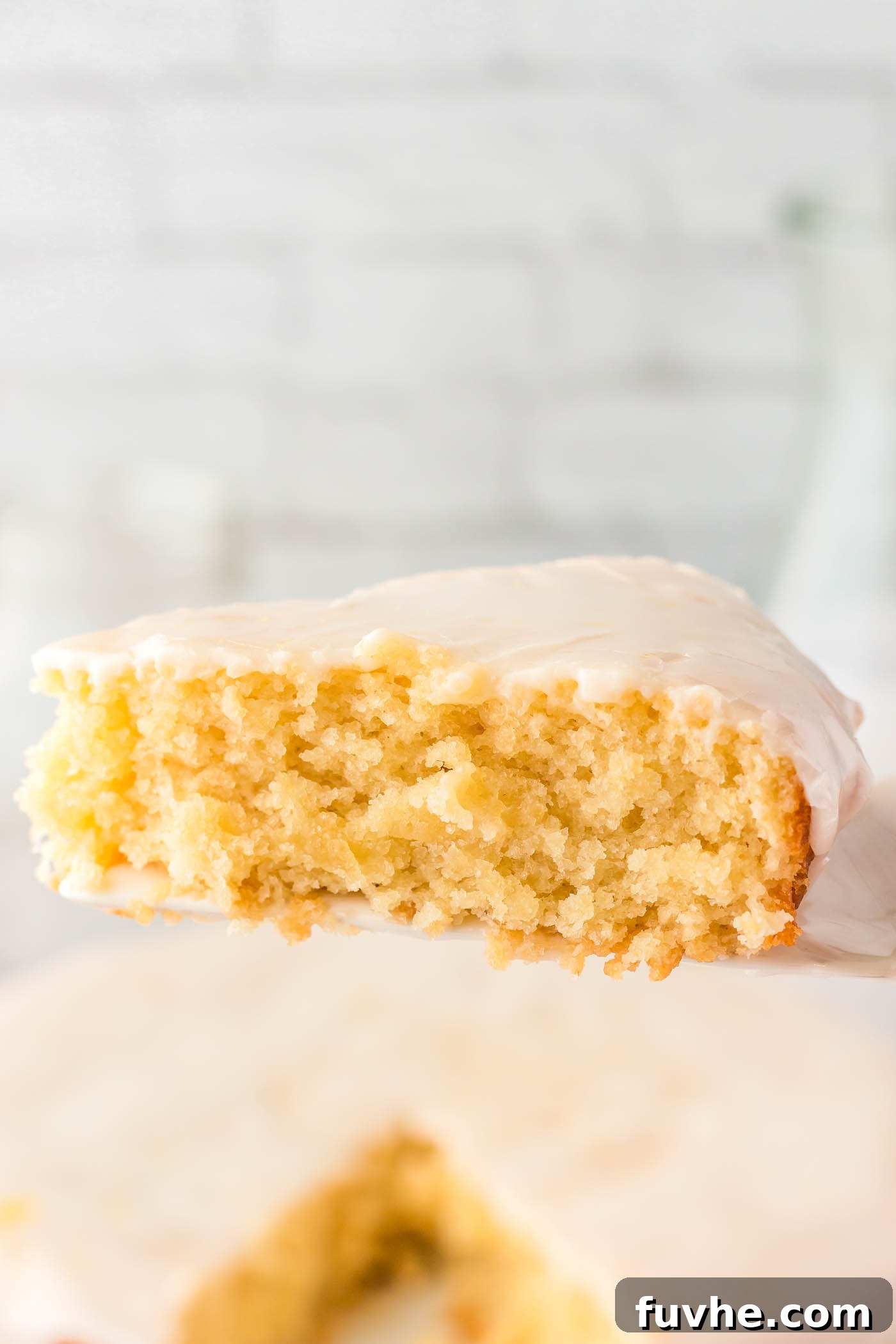 A perfectly baked slice of vegan olive oil cake, elegantly topped with a delicate powdered sugar glaze, is held on a spatula above the pristine whole cake.