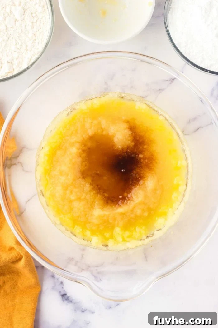 Vanilla extract and unsweetened applesauce are added to the creamed olive oil and sugar mixture in a large glass bowl on a marbled countertop.