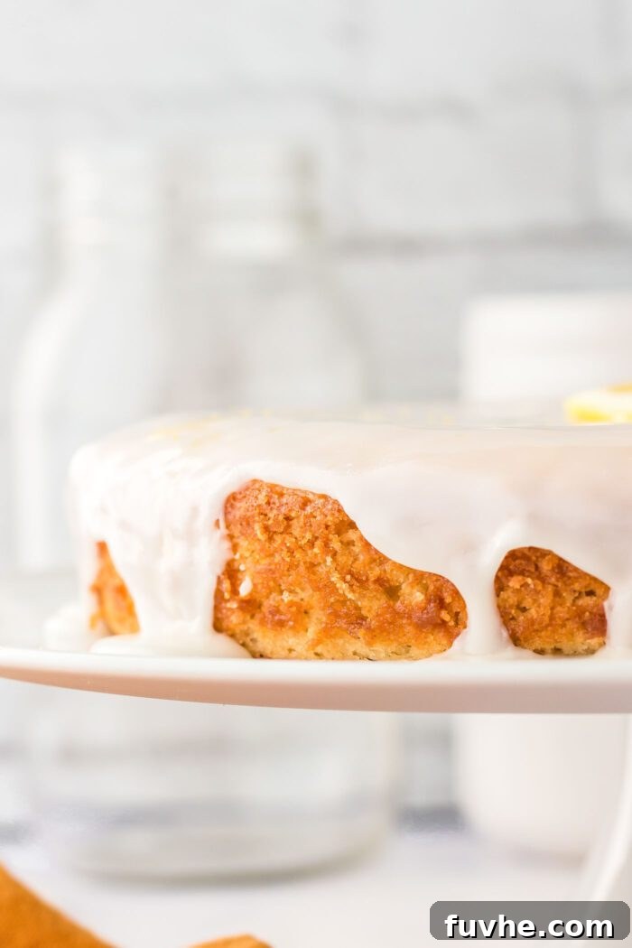 A beautifully presented vegan olive oil cake, topped with a luscious powdered sugar glaze and garnished with a fresh lemon slice, sits elegantly on a cake stand with blurred background elements.