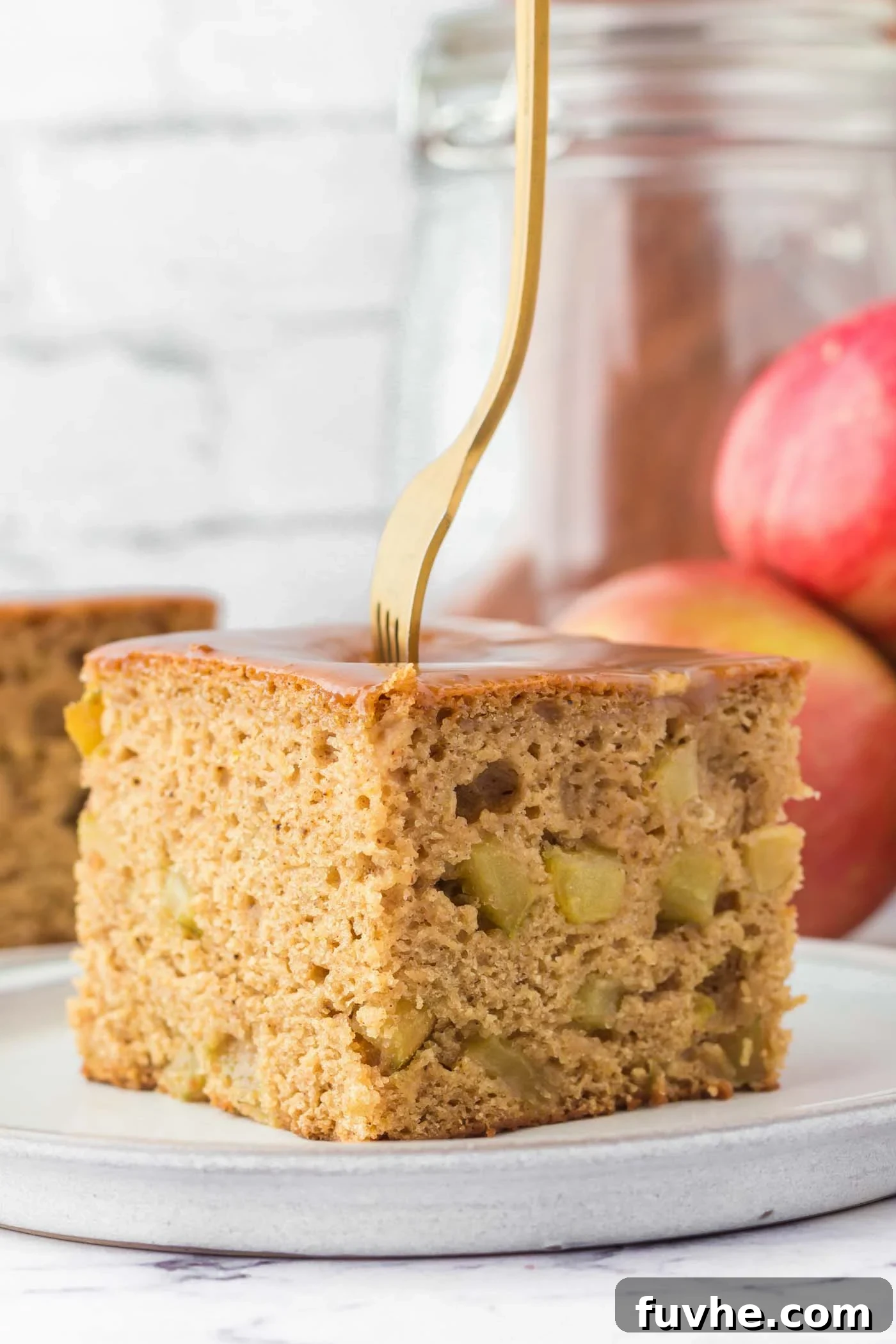 A large square of vegan apple cake on a plate. The cake is topped with a caramel glaze, there's a fork stuck in the cake and you can see some pieces of diced apple in the cake.