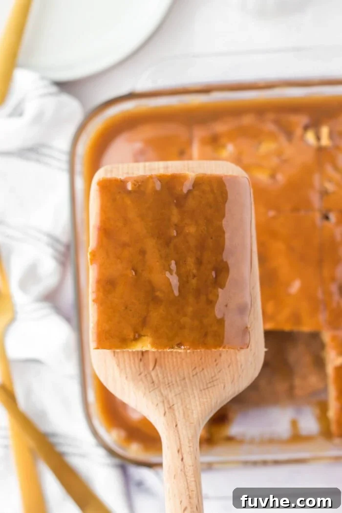 A large wooden spatula with a square of caramel glazed apple cake on it held over the cake it was removed from in a glass baking dish.
