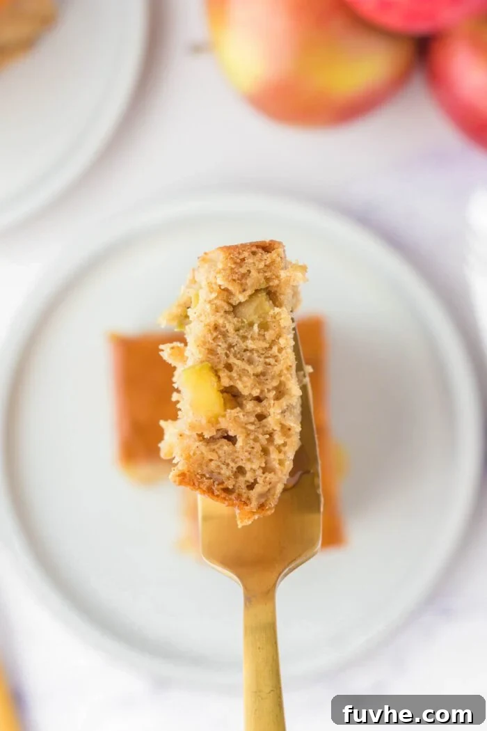 A fork with a piece of caramel apple cake on it held over a square of apple cake on a small plate.