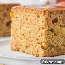 Close up of a large square of apple spice cake from an angled view on a small plate. You can see bits of apple in the cake and it's topped with a thin layer of caramel.