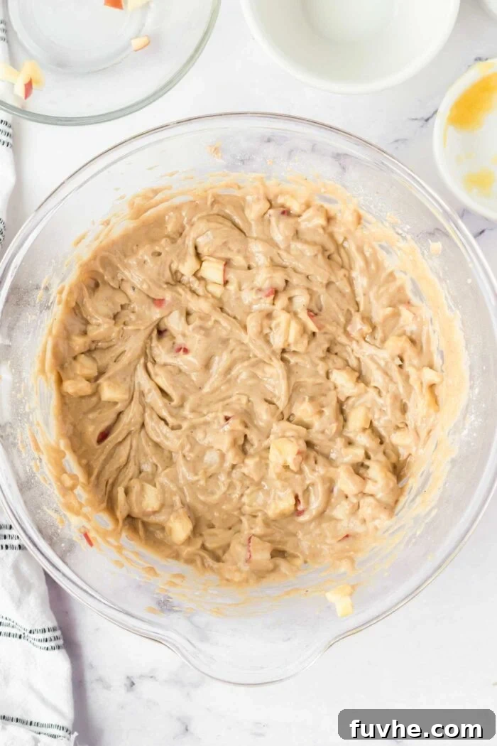 Raw apple cake batter with pieces of diced apple in a large glass mixing bowl.