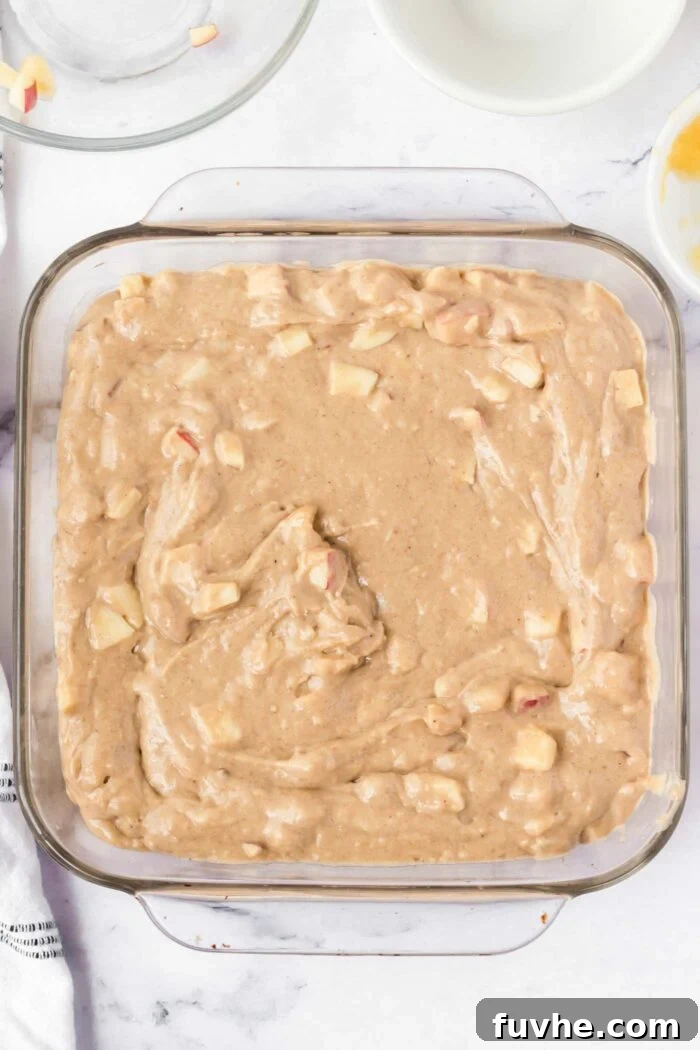 Raw apple cake batter with pieces of apple in it in a square glass baking dish.