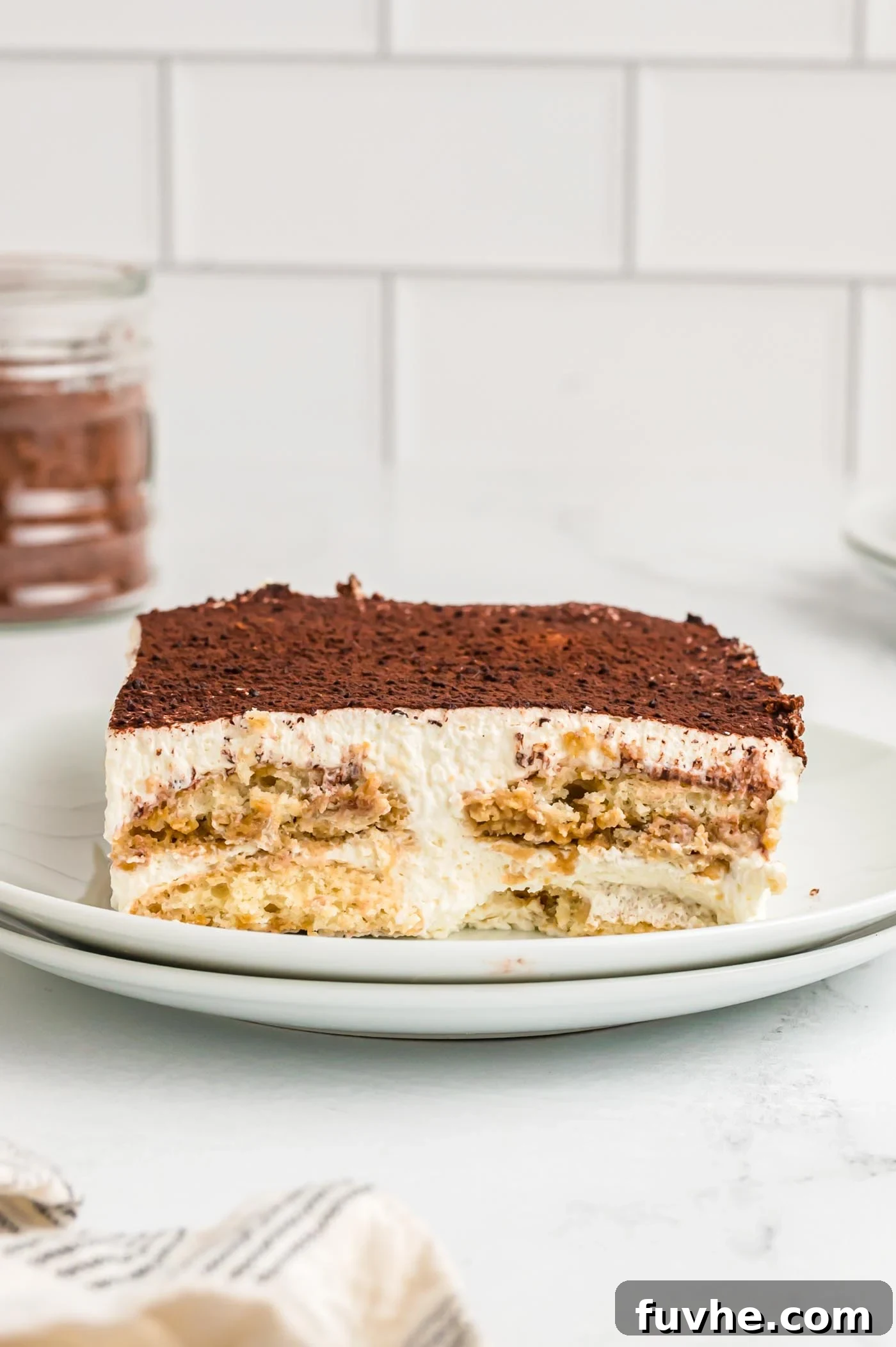 Decadent Vegan Tiramisu 2 A square serving of tiramisu cake on a plate. You can see the layers of ladyfingers, marscapone cream filling and cocoa powder on top.