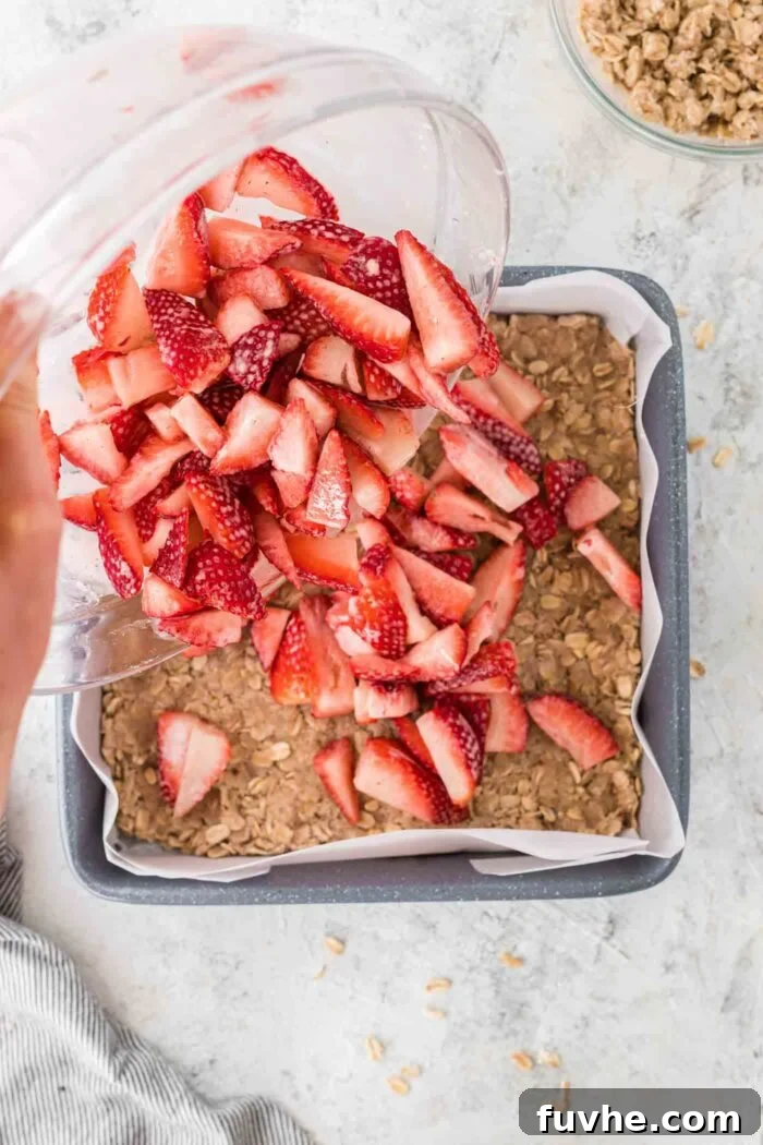 Irresistible Vegan Strawberry Crumble Bars 6 Diced fresh strawberries, coated in tapioca starch and lemon juice, being poured from a glass bowl onto the pressed oatmeal crust in a square baking pan.
