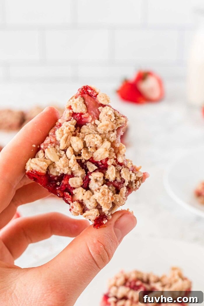 Irresistible Vegan Strawberry Crumble Bars 10 A hand holding a freshly baked vegan strawberry crumble bar with a bite taken out, revealing the vibrant strawberry filling and crumbly oat layers. More bars and fresh strawberries are blurred in the background.