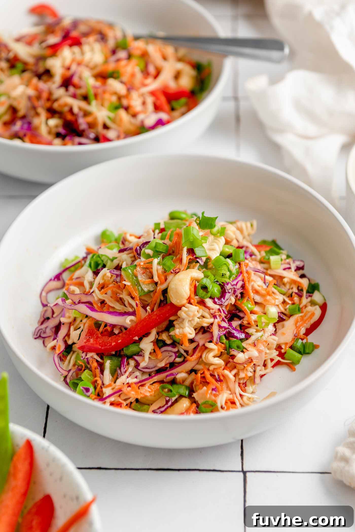 A beautifully presented bowl of crunchy Asian ramen noodle salad, garnished with cashews, edamame, and a colorful array of vegetables like shredded cabbage, carrots, green onions, and bell pepper. Another bowl is subtly blurred in the background, suggesting a generous serving.
