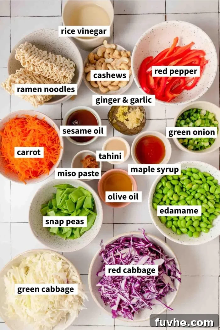 Individual bowls holding various ingredients for a ramen noodle salad, including shredded cabbage, grated carrots, chopped bell pepper, edamame, and cashews, ready for assembly. Each ingredient is clearly visible and separated.