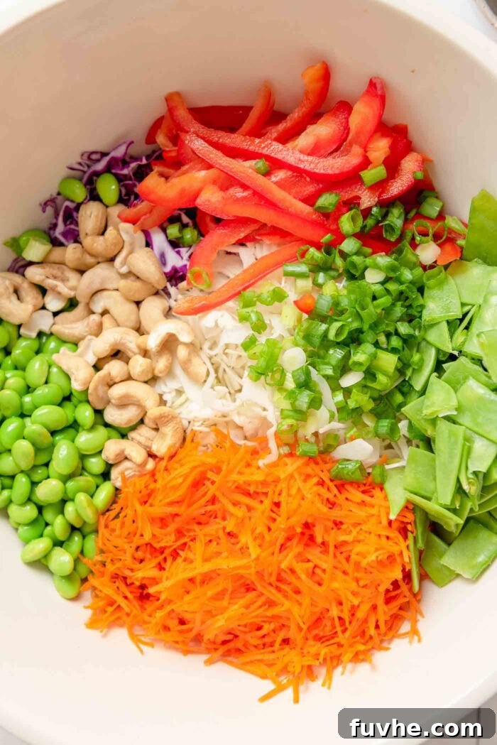 A large ceramic mixing bowl filled with a colorful array of prepared salad ingredients: grated carrot, chopped snow peas, red bell pepper, roasted cashews, edamame, shredded red cabbage, and green cabbage, ready to be dressed.