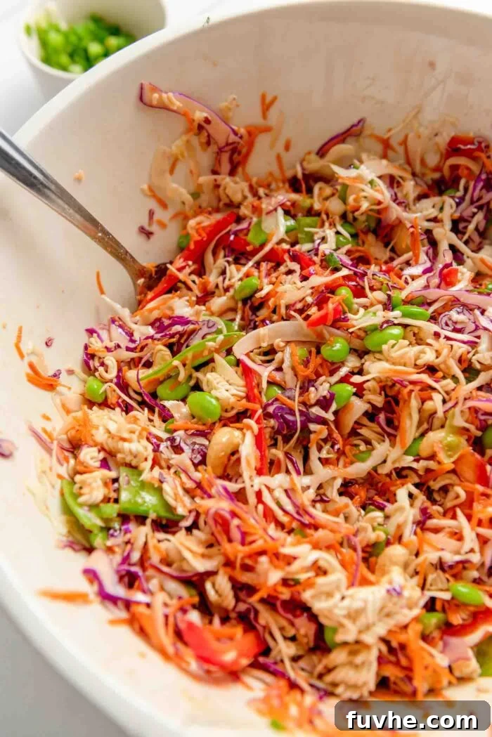 A vibrant ramen noodle salad, featuring shredded red and green cabbage, cashews, edamame, and carrots, perfectly mixed in a large bowl with a metal serving spoon nestled within. The noodles are evenly coated in the dressing.