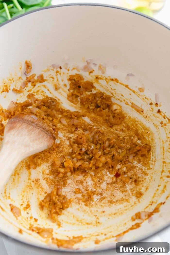 Minced shallot, garlic, grated ginger, and fragrant curry powder cooking together in a large soup pot with a wooden spoon, creating an intensely aromatic paste.