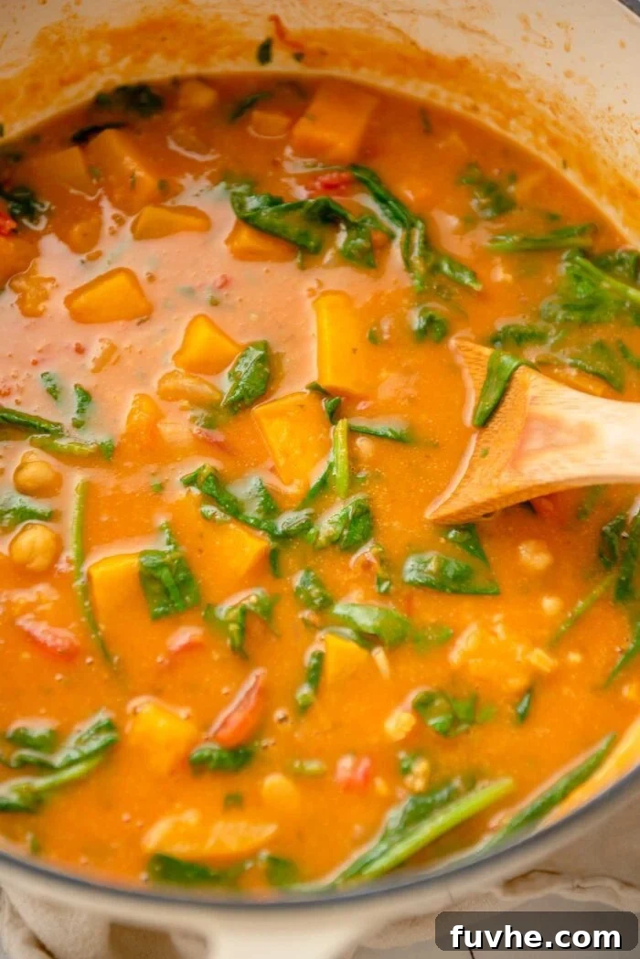 A large pot of vibrant butternut squash curry, with fresh spinach visibly wilting into the creamy sauce, accompanied by hearty chickpeas. A wooden spoon rests inside, ready to serve.
