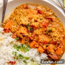 Overhead view of a sweet potato and red lentil curry in a bowl with rice and a wedge of lime. A spoon rests in the bowl and there are two more small spoons beside the bowl on the counter.